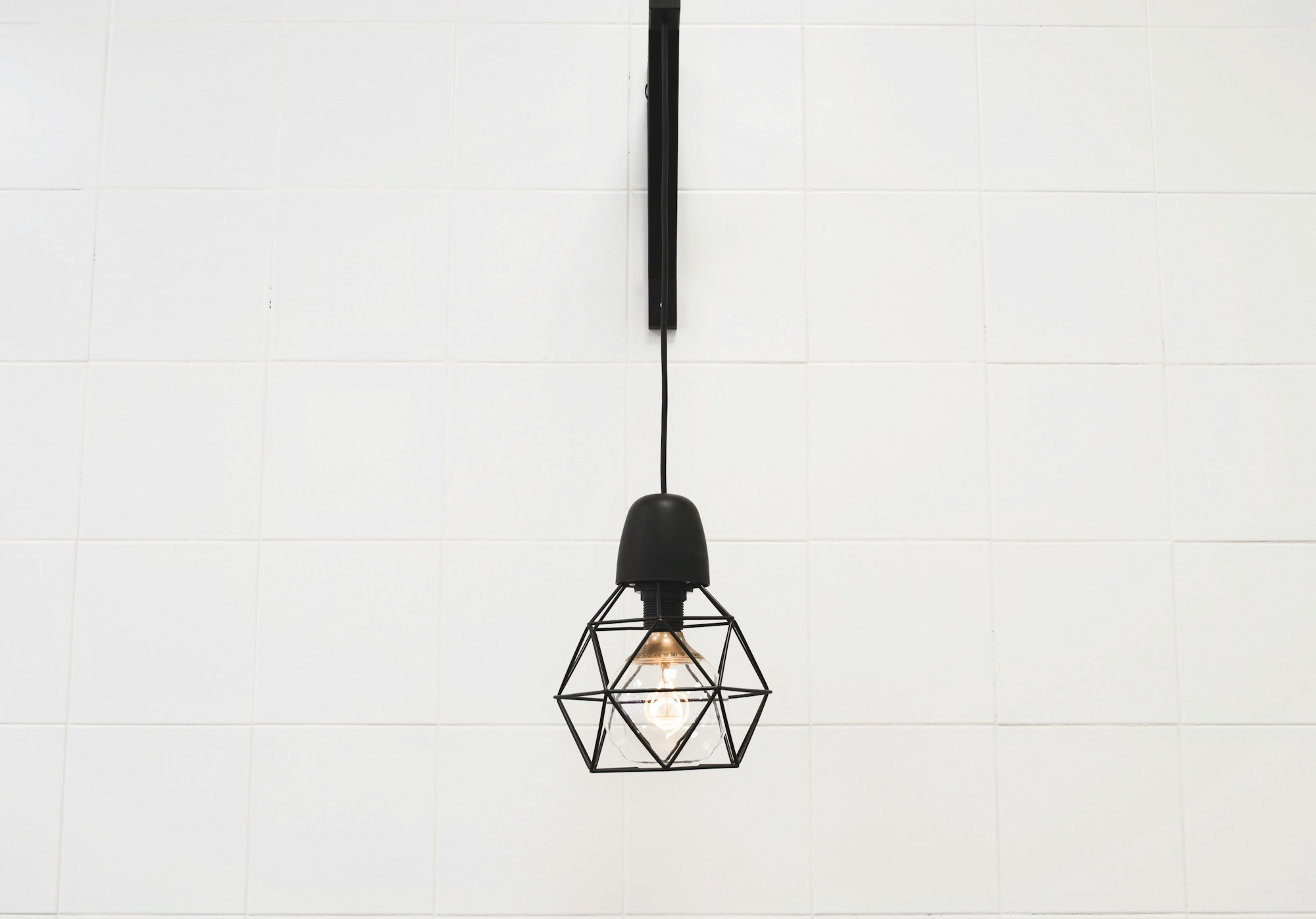 A Vigorous wire-cage pendant, lit, against a bright tiled wall.
